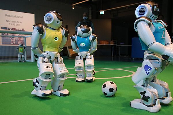 drei Roboterfußballer auf einem Kunstrasenspielfeld in einem Raum, ein Ball liegt in der Mitte von ihnen