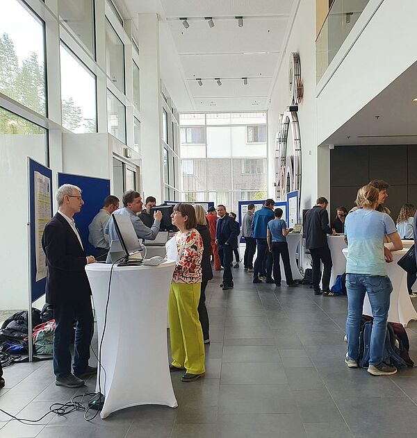 Menschen stehen in einem Foyer zusammen und unterhalten sich. Es sind Monitore und Pinnwände mit Postern zu sehen.