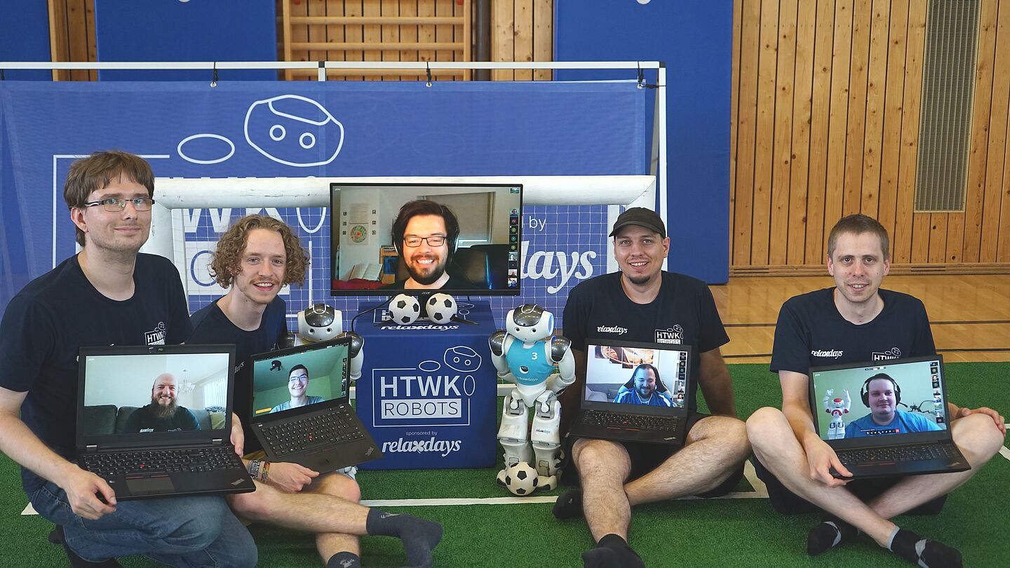 Teammitglieder (in Präsenz, v.l.n.r.): Tobias Jagla, Felix Loos, Stephan Bischoff und Jörg Schließer. Auf den Laptops zu sehen sind Teammitglieder, die von Ferne unterstützt haben (v.l.n.r.): Teamchef Rico Tilgner, Erik Behrendt, Tobias Kalbitz (groß Vier Männer mit Laptops auf dem Schoß vor kleinem Tor und mit humanoidem Roboter