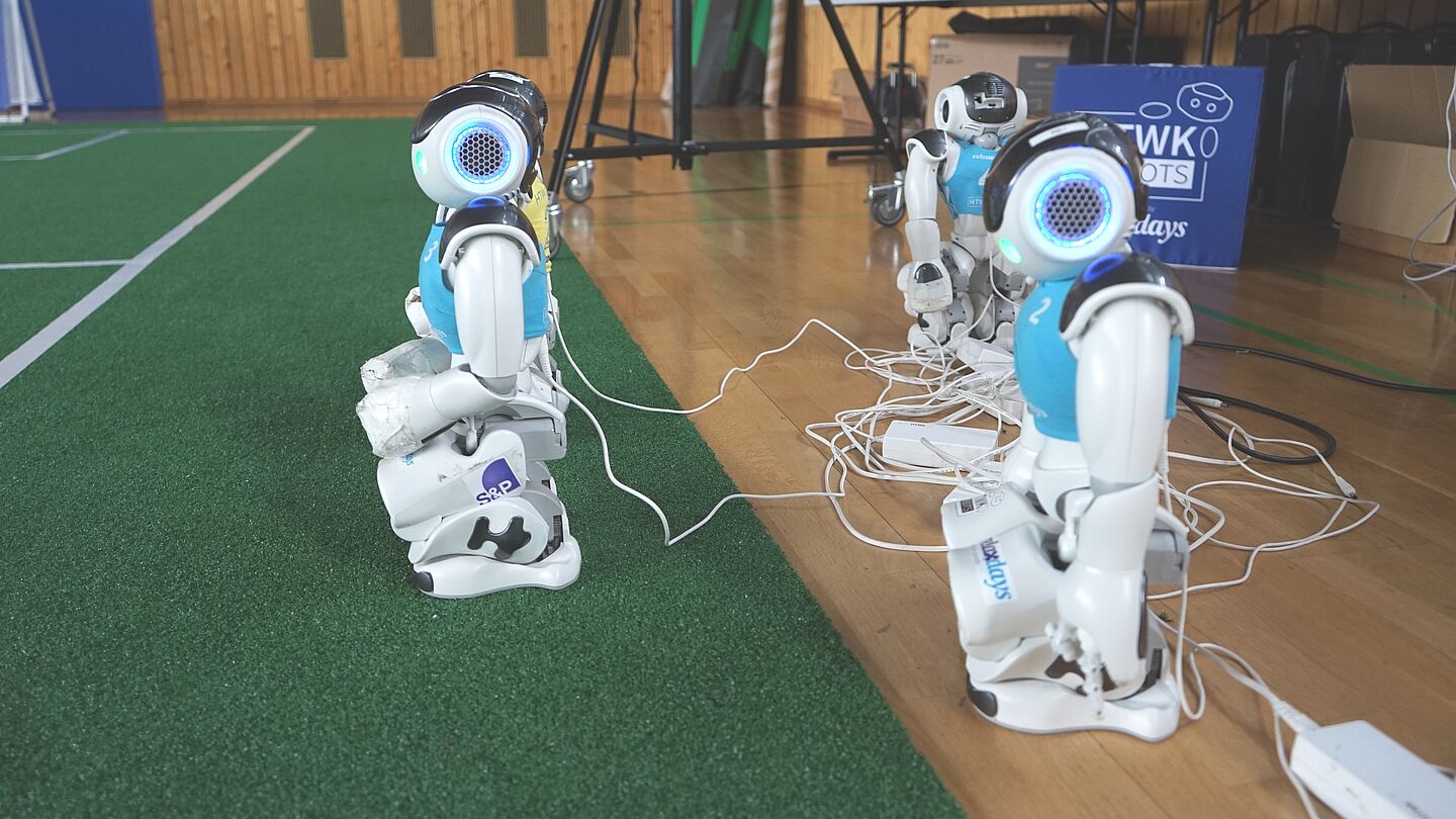 Auch Naos müssen ihr Akku laden. (Foto: Pressestelle/HTWK Leipzig) drei humanoide Fußball-Roboter am Ladekabel
